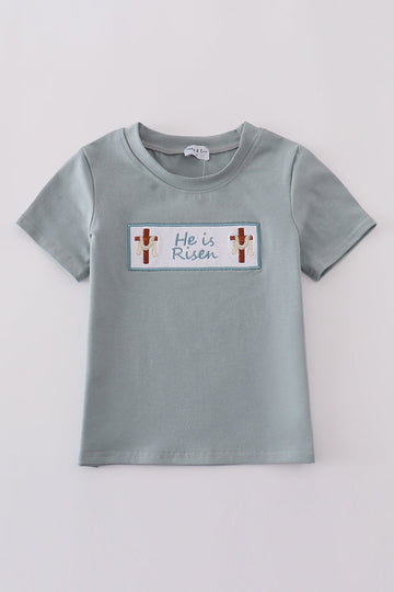 Blue children's top with "He is Risen" embroidered in white script.